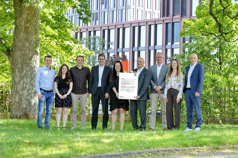 Gruppe von neun Personen steht auf einer Wiese vor einem modernen Bürogebäude, eine Person hält eine Urkunde